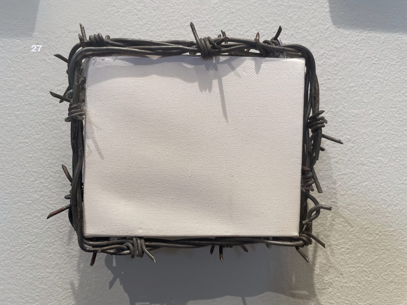A small, completely blank white canvas surrounded by a frame of barbed wire